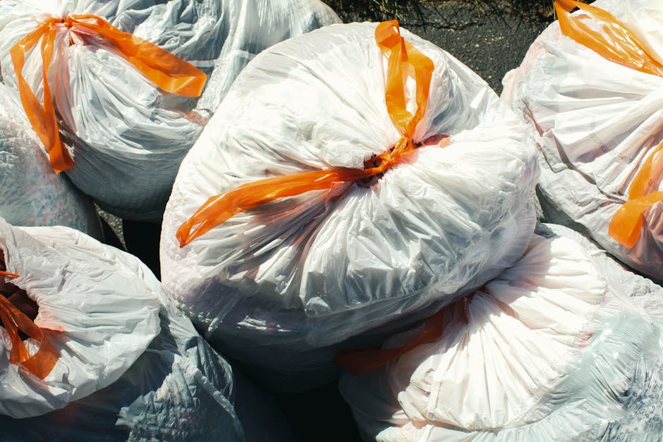 A collection of multiple large white plastic rubbish bags filled with waste, each tightly knotted at the top with orange plastic ties, placed outdoors on a paved surface. The bags appear to be full an